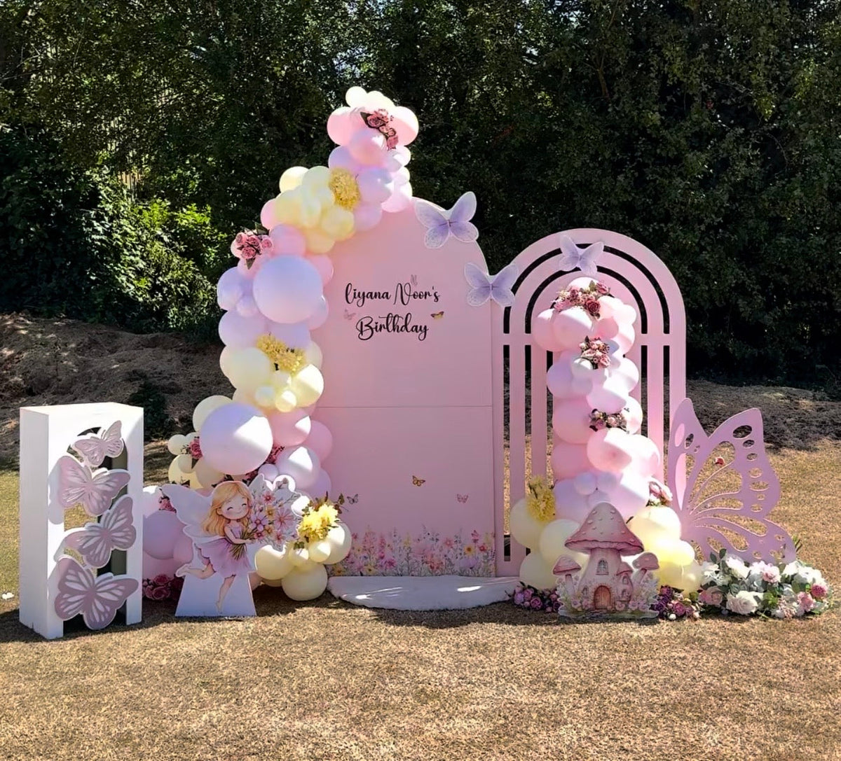 Bespoke fairy garden birthday display with personalised arch backdrop, butterfly cutouts and a mushroom house styled with a pastel balloon garland
