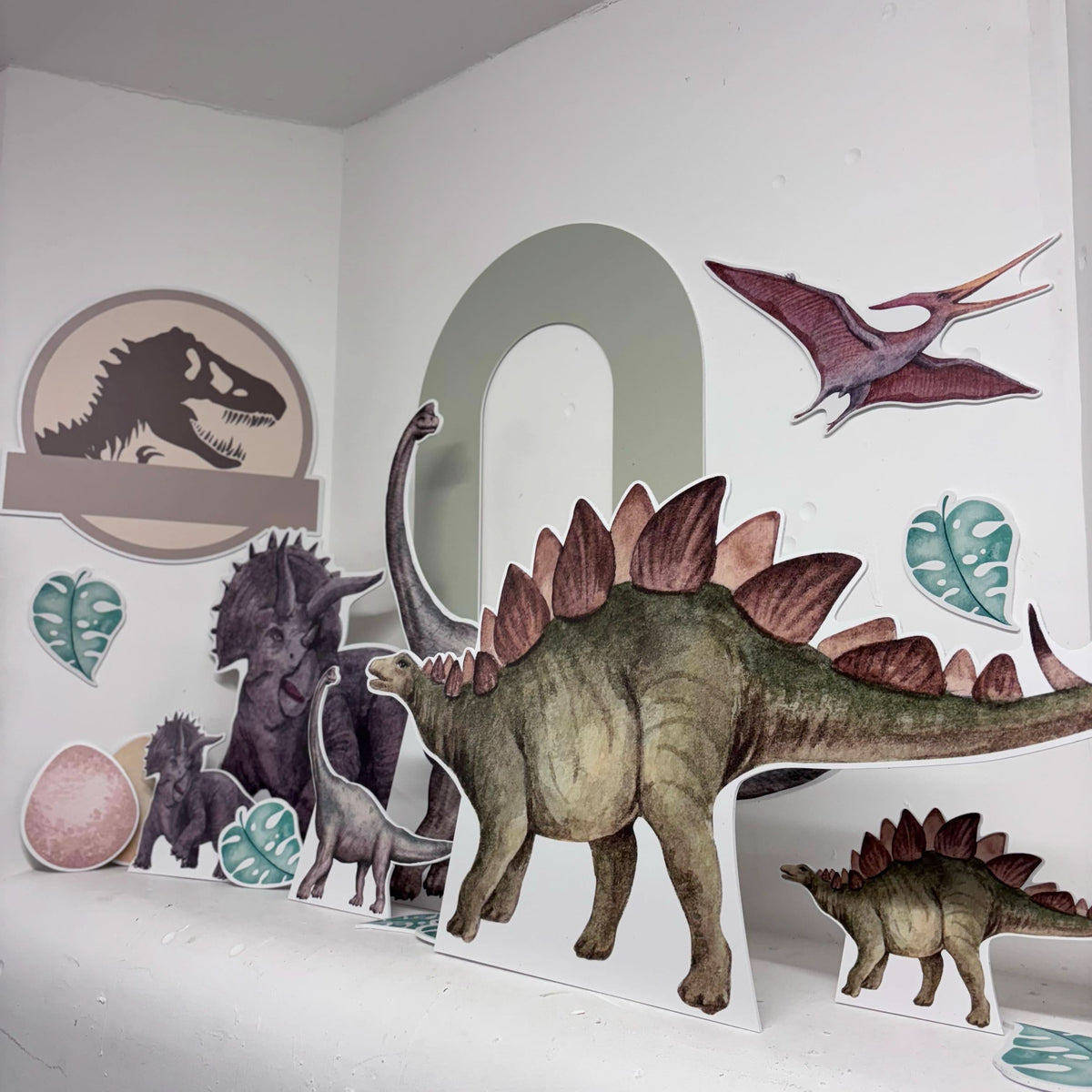 Close-up of Dinosaur World waterproof foamex cutouts showing stegosaurus, brachiosaurus, pterodactyl, and T-rex logo with green arch sail board and tropical leaf decorations