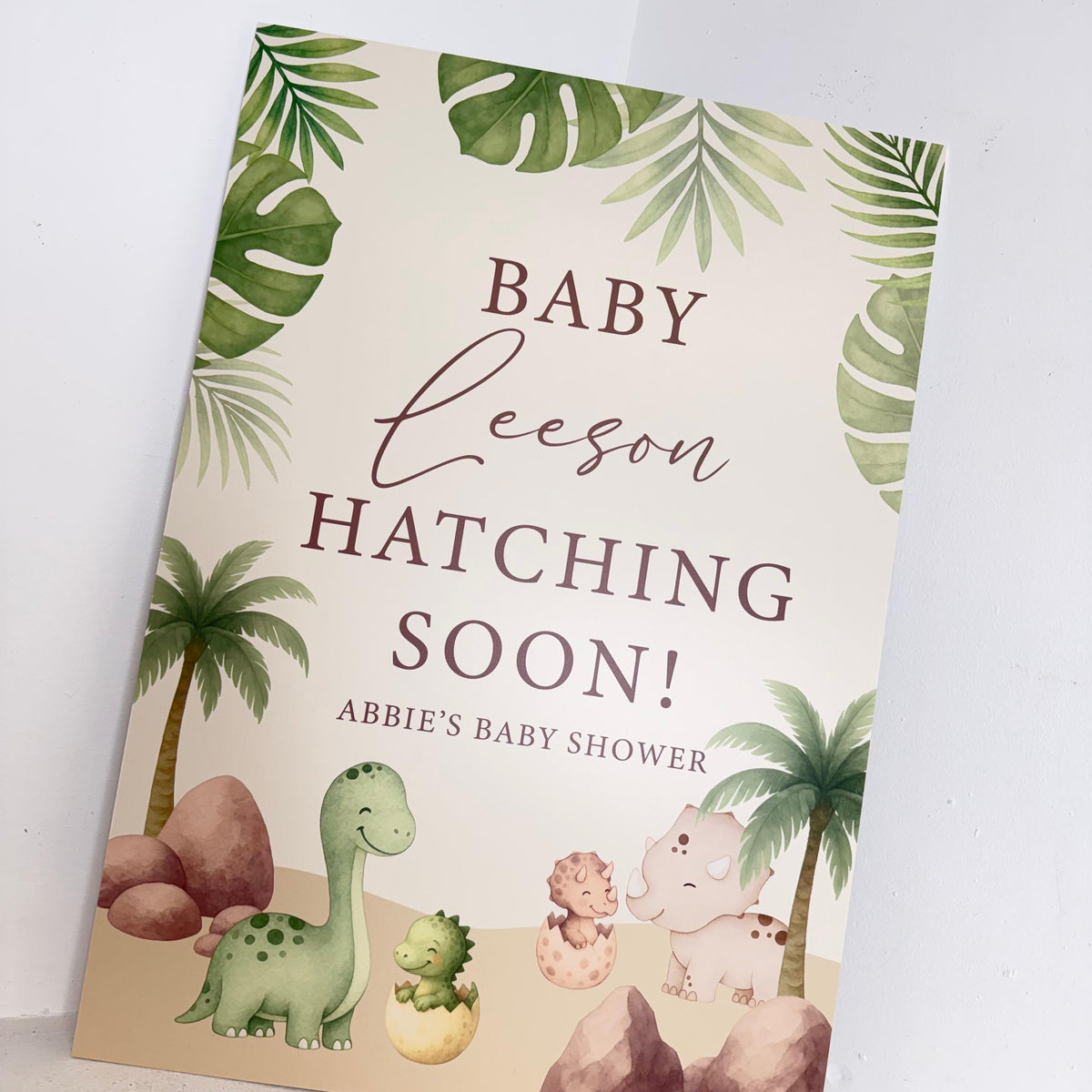 Close-up angled view of Baby Hatching dinosaur themed baby shower foamex easel sign with personalised name and tropical leaf design