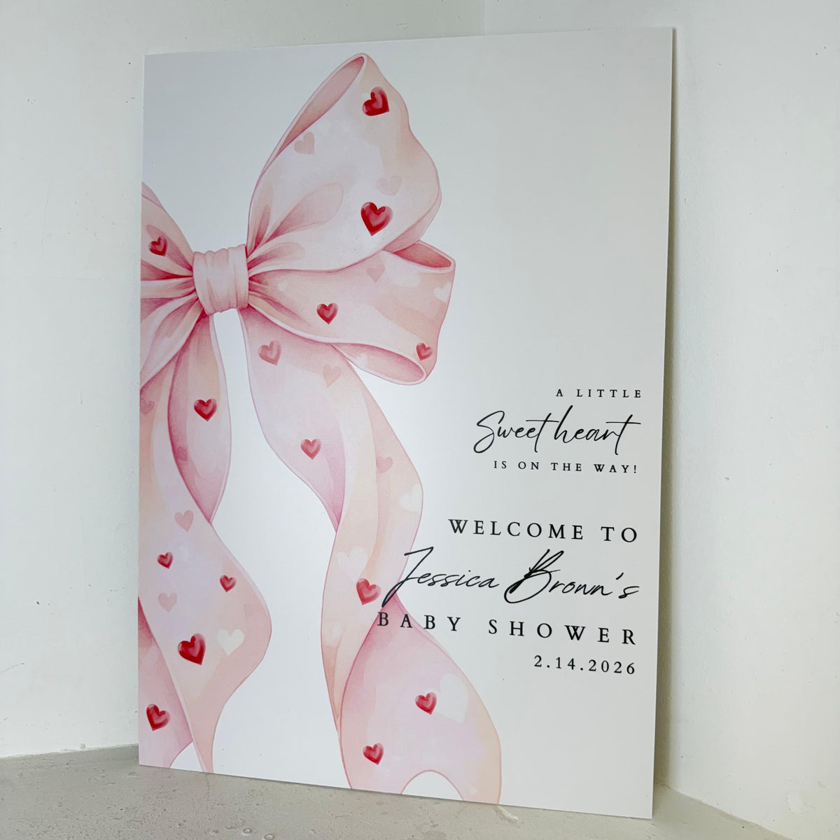 Close-up angled view of Sweetheart On The Way baby shower foamex easel sign showing personalised name and date with pink ribbon detail