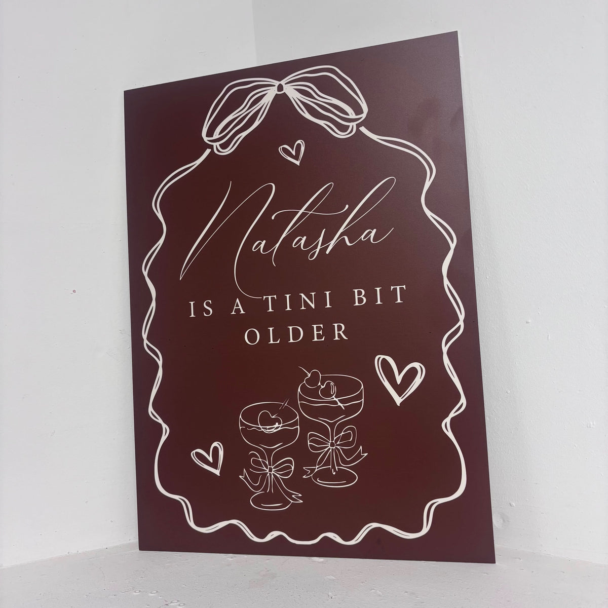 Tini Bit Older easel sign angled view showing burgundy design with cocktail illustrations and personalised name on freestanding foamex board